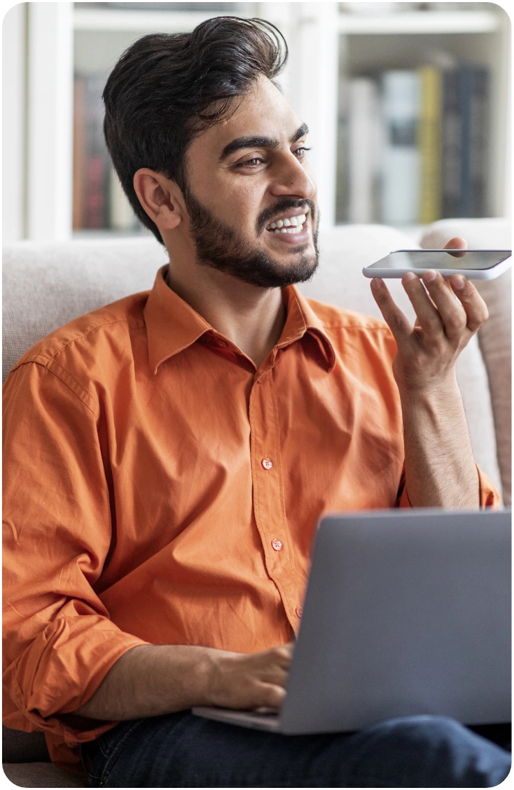 Man in an orange shirt speaking into a smartphone while using a laptop.